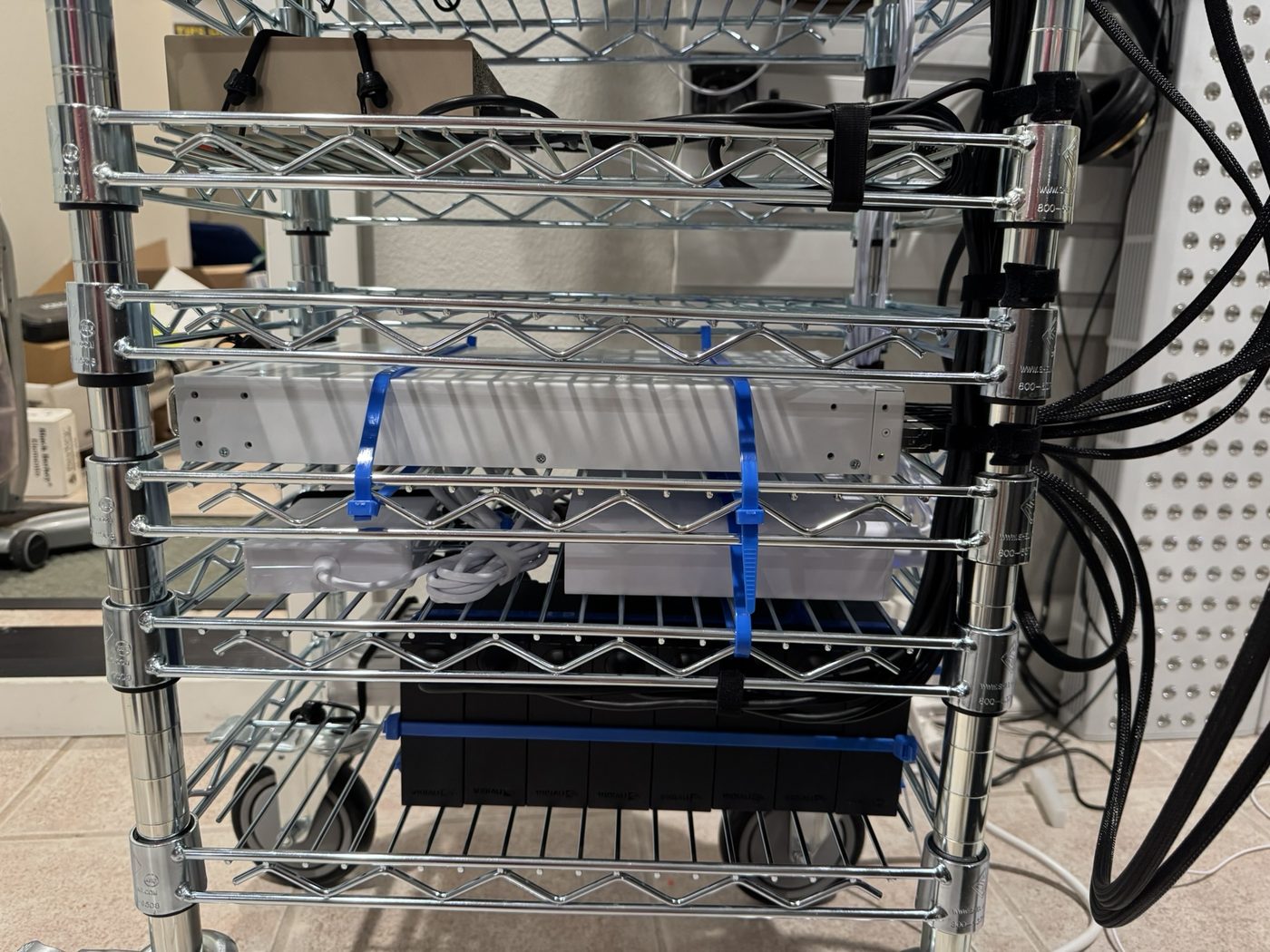 The lower decks of GhettoRak: a white switch zip-tied to a wire shelf, power bricks with coiled white cords beneath it, and a black unit on the deck below.