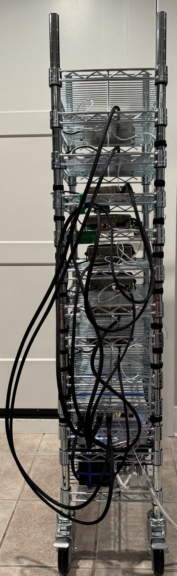 GhettoRak from the rear: heavy black braided cables strapped to both uprights drape in large loops in front of the stacked units, with white patch cords, a white switch with status LEDs, and power bricks on the lower shelves.