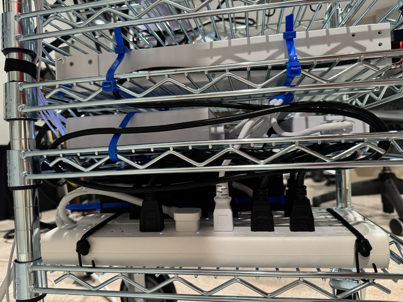 A white Tapo power strip tied along the front of a lower wire shelf, black and white plugs in several outlets, small blue LEDs lit, cable bundles and blue zip ties around it.